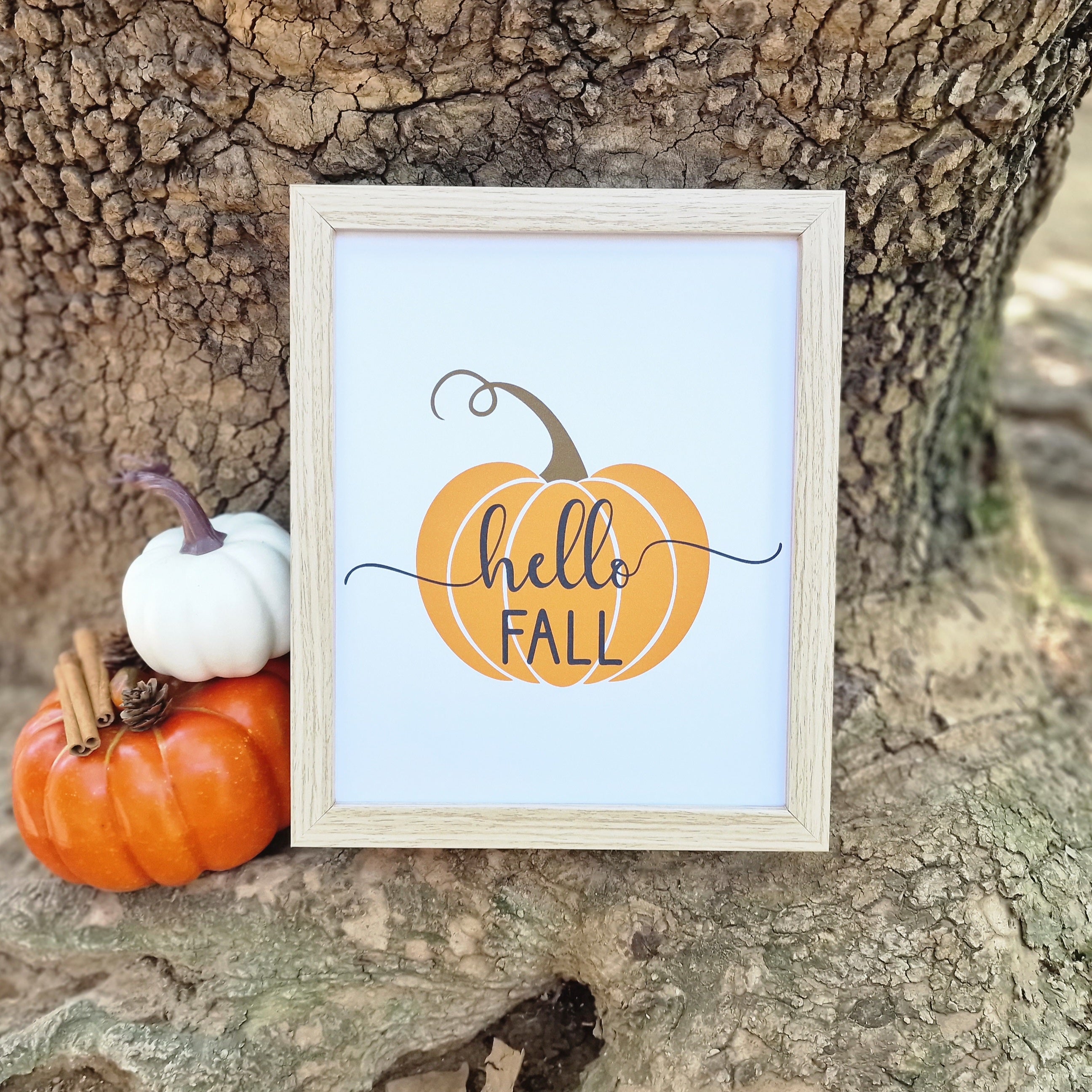 Hello Fall πόστερ τοίχου με ξύλινο πλαίσιο με ατάκα πάνω σε σχέδιο κολοκύθας. Η κορνίζα βρίσκεται πάνω σε κορμό δέντρου δίπλα σε πορτοκαλί και άσπρη κολοκύθα.